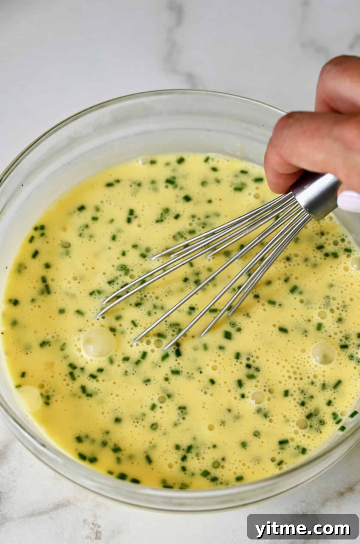 A glass bowl holds whisked eggs and finely chopped chives, part of the creamy quiche filling.