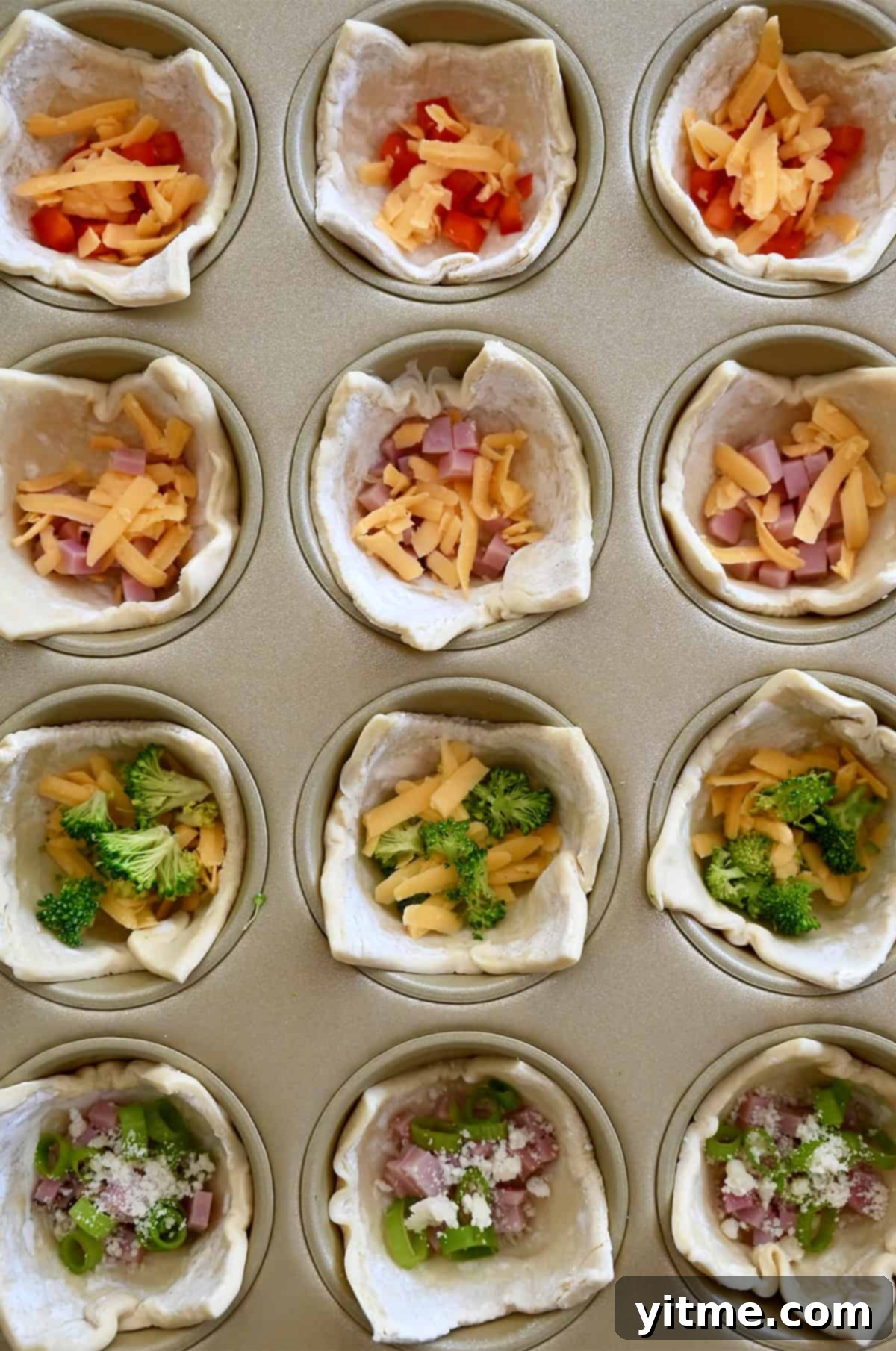 Puff pastry cups in a muffin tin, each filled with a different combination of ingredients like diced ham and cheese, broccoli, or red pepper, ready for the egg mixture.