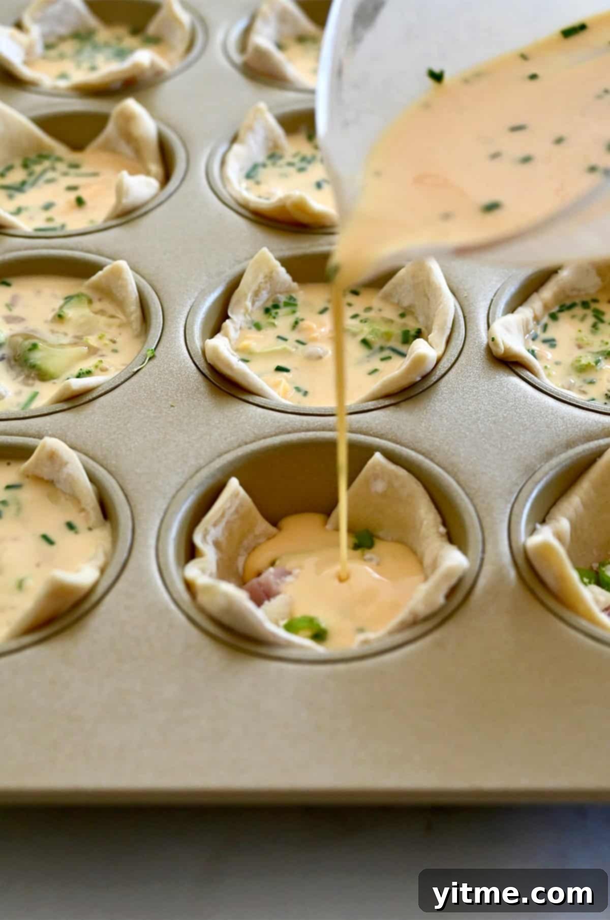 The process of pouring whisked egg mixture into puff pastry cups, already filled with various ingredients, in a muffin tin.