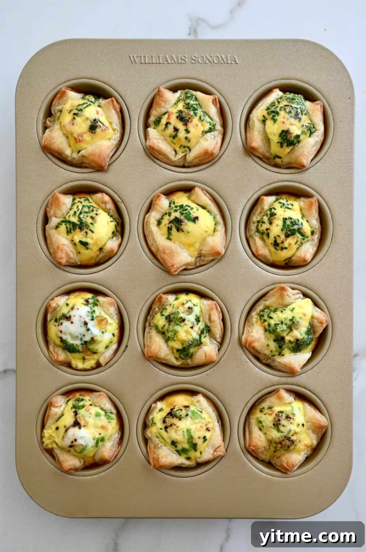 A muffin tin filled with freshly baked, golden-brown mini quiches with puff pastry crusts.