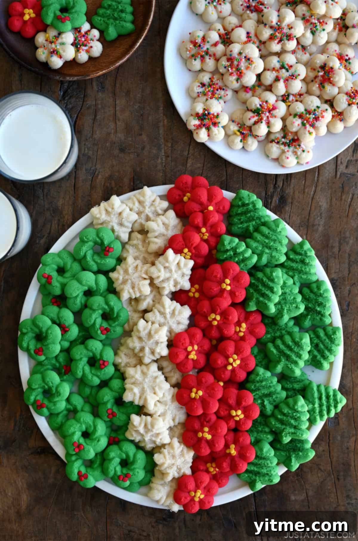 A festive assortment of traditional spritz cookies, including green Christmas trees, red rosettes, sprinkle-adorned snowflakes, and verdant wreaths on a cookie tray.