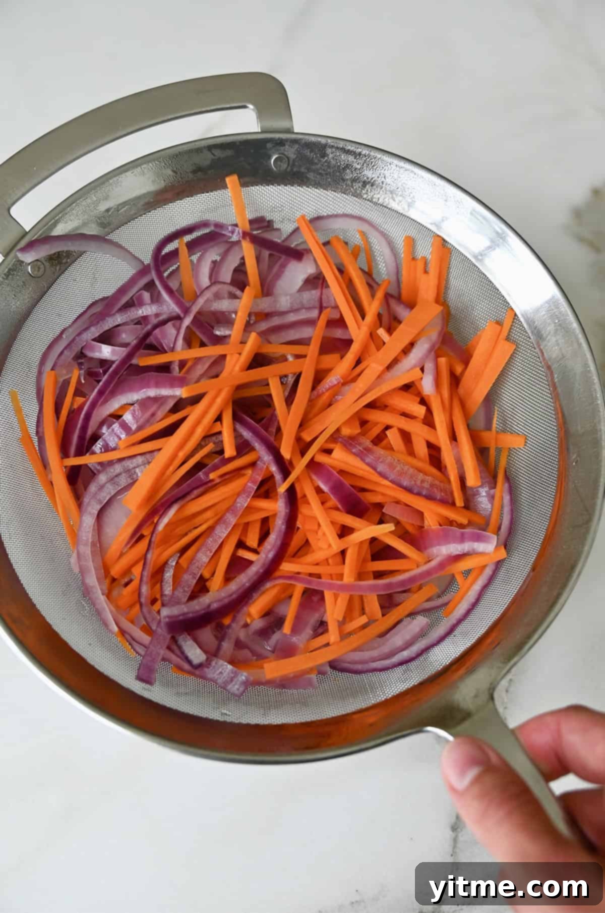 A hand holds a fine mesh strainer filled with pieces of red onion and carrots, just blanched and drained, ready for the pickling brine.