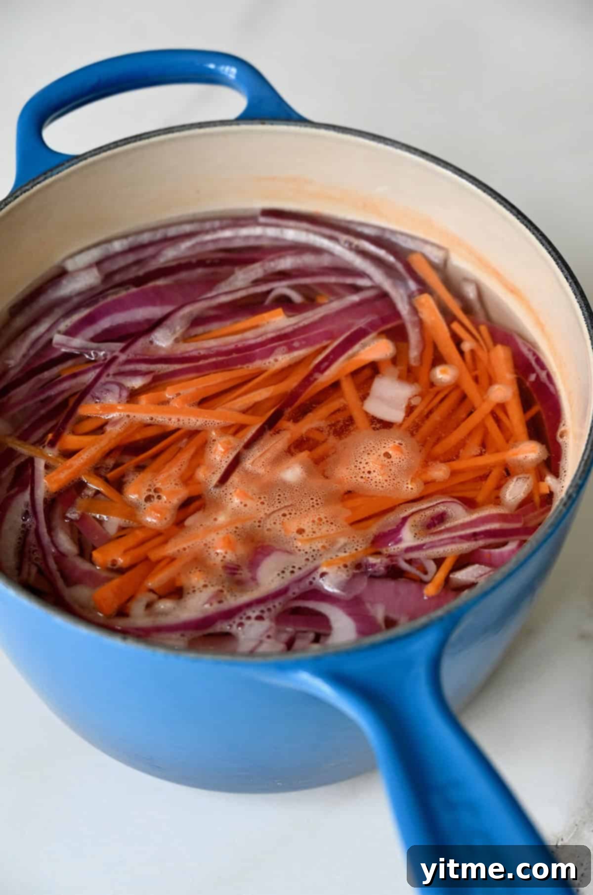 A blue saucepan is filled with sliced red onions, diced carrots, and the pickling liquid, simmering together on the stovetop.