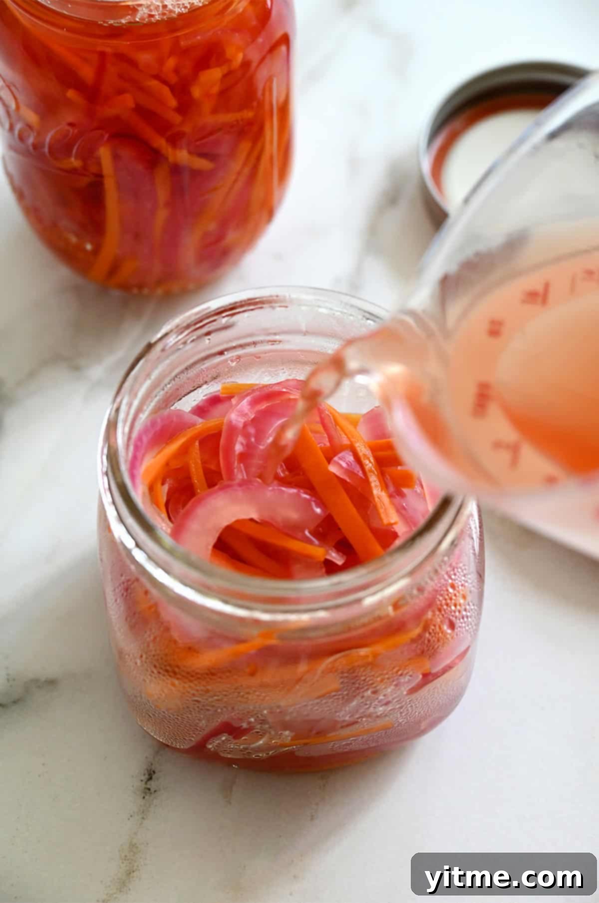 Pickling mixture is being poured from a liquid measuring cup into a glass jar already filled with red onions and carrots. A second filled jar and a lid sit nearby on a white marble surface.