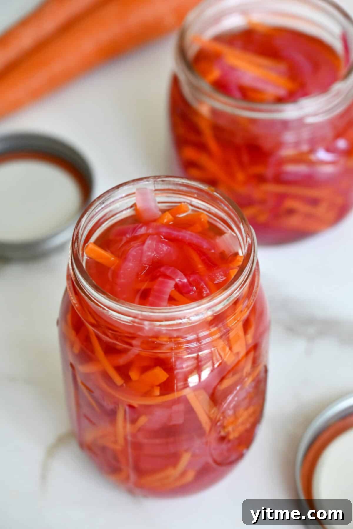 Two glass jars are filled with pickled red onions and carrots. The lids and some carrots sit nearby.