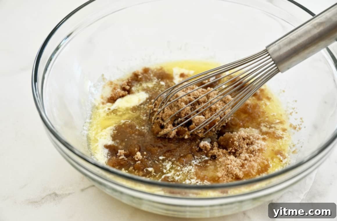 A whisk vigorously mixing melted butter and brown sugar in a clear glass bowl, showcasing the rich, amber color of the mixture.