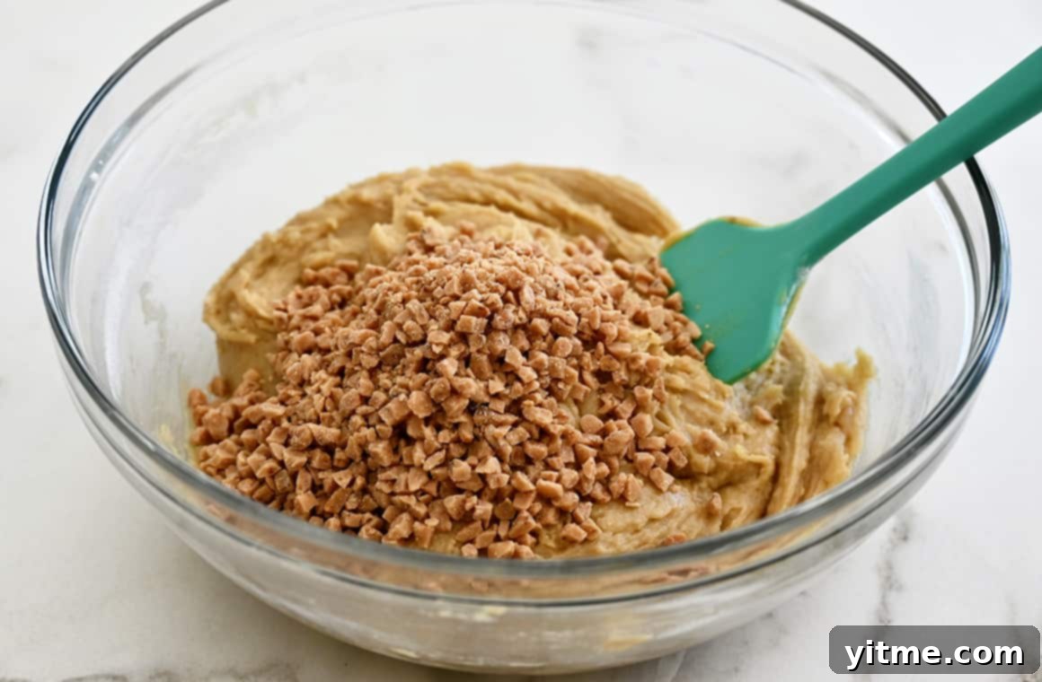 A vibrant teal spatula gently folding blondie batter with scattered toffee bits in a clear mixing bowl, highlighting the rich texture.
