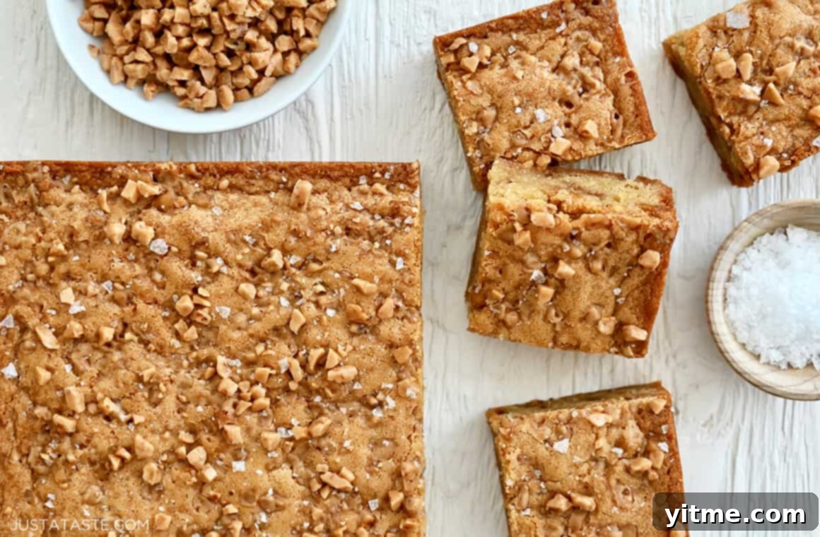 A top-down view of perfectly sliced toffee blondies arranged next to small decorative bowls holding glistening sea salt flakes and crunchy toffee bits, ready for serving.
