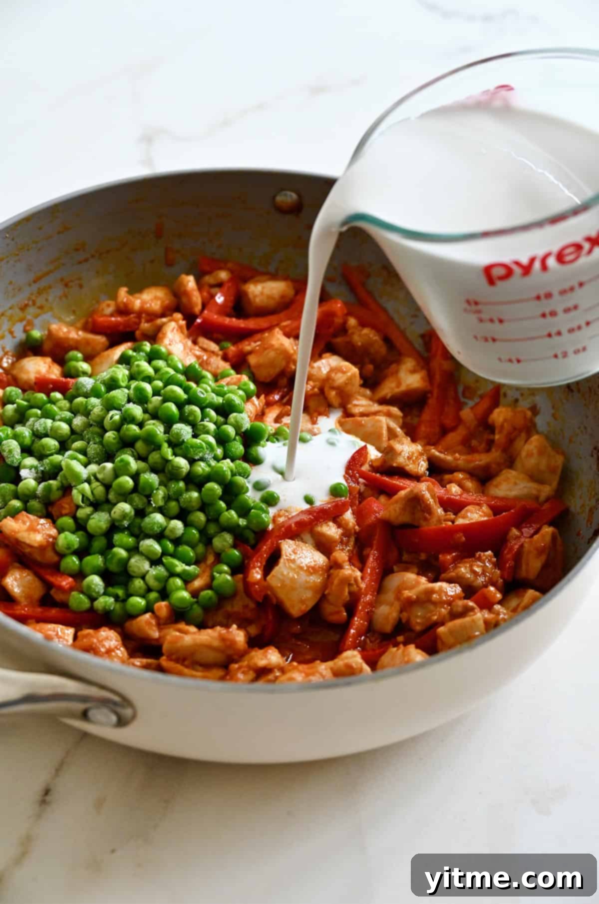 Coconut milk being poured into a skillet with frozen peas, cooked chicken and sautéed veggies.