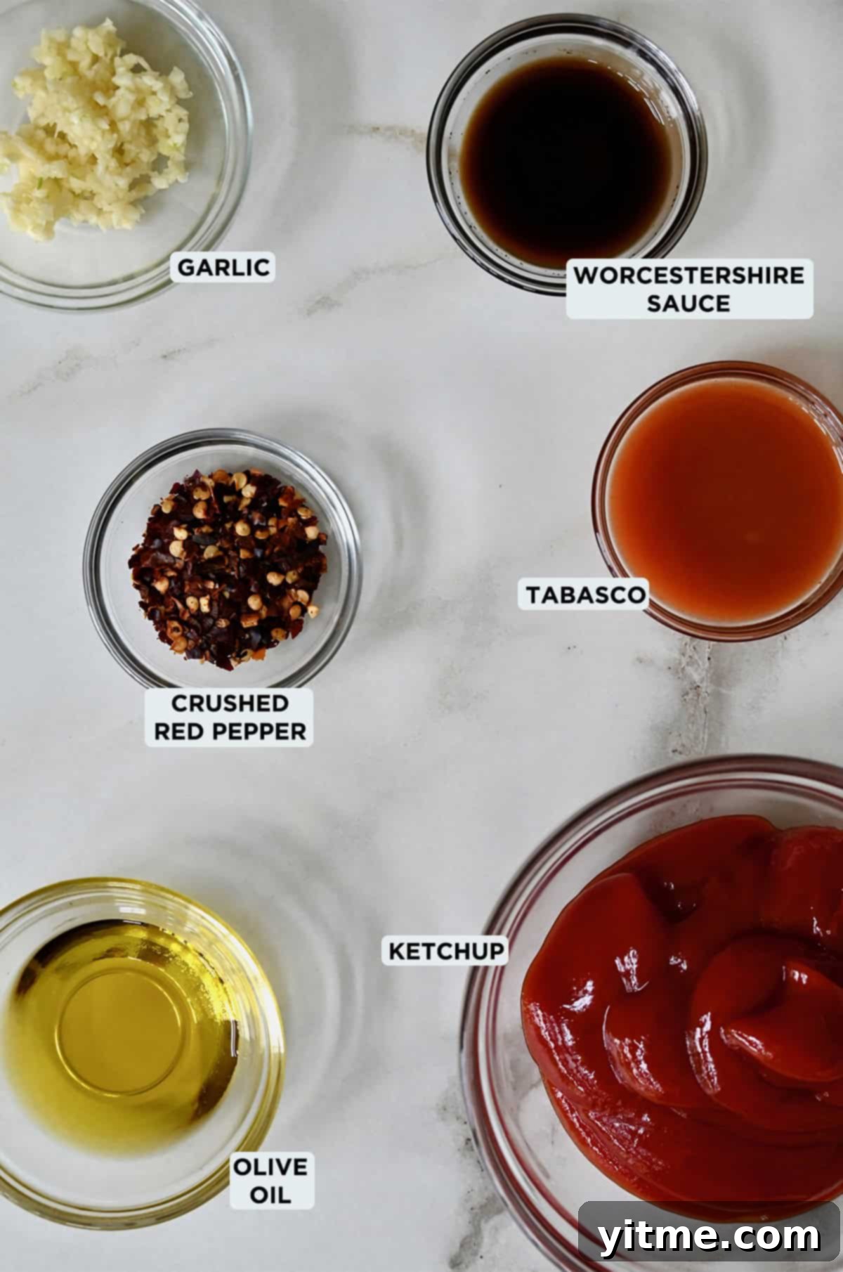 Small glass bowls holding ingredients for spicy ketchup: ketchup, olive oil, minced garlic, crushed red pepper, Tabasco.