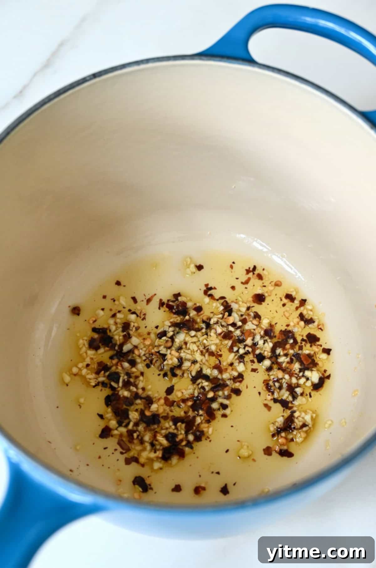 Garlic and red pepper flakes being sauteed in oil in a blue enamel cast iron saucepan.