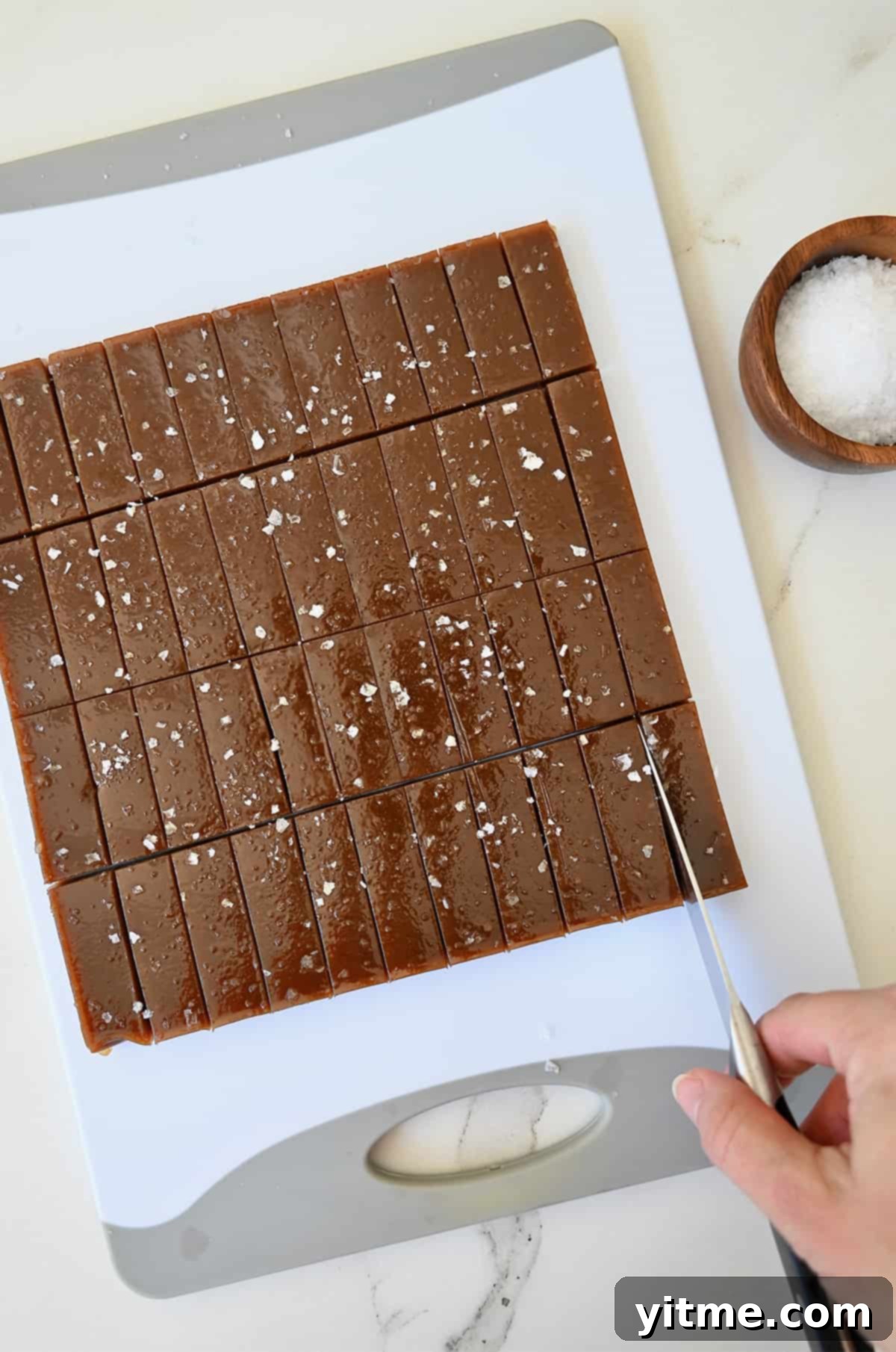 A hand holding a knife cuts into a block of soft caramels on a cutting board.